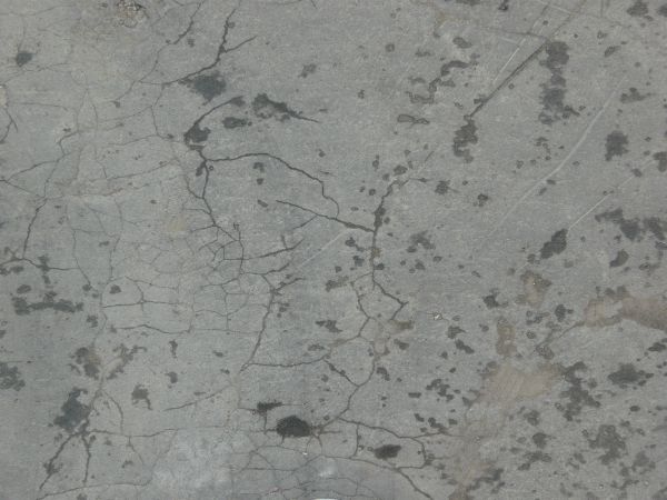 Grey asphalt texture, covered with cracks of various sizes and depths, and many shallow chipped holes.